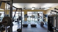 This fitness center is thoughtfully designed to provide a comprehensive workout environment for users of all fitness levels. The space is anchored by a row of modern cardio machines, including treadmills and ellipticals, all positioned to face a generous wall of large floor-to-ceiling windows that flood the room with natural light and offer pleasant views of the surrounding outdoor landscaping and building exterior. The bright, open atmosphere is enhanced by warm yellow accent walls that lend an energetic ambiance to the space. Overhead, multiple ceiling fans with integrated lighting fixtures provide consistent airflow and comfort during workouts. To one side, a dedicated strength training area features a cable machine and free weight equipment, with mirrored walls that visually expand the space and allow users to monitor their form. A stability ball and additional equipment are neatly arranged throughout, making efficient use of the available floor area. The durable, dark rubberized flooring provides both safety and functionality across the entire space. Overall, the fitness center strikes a balance between practicality and comfort, offering a well-lit, ventilated, and fully equipped environment suited for a wide range of exercise routines.