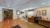 This single-floor facility is designed to offer a well-rounded communal experience through a series of thoughtfully furnished and connected spaces. The entry area makes an immediate impression with its striking reddish-orange marbled flooring, which extends through the main corridors and transitions seamlessly into carpeted lounge and activity areas. Recessed lighting throughout, set within drop tile ceilings, ensures the entire floor is evenly and warmly illuminated.

The central lobby and lounge areas feature multiple seating arrangements with plush sofas, accent chairs, and glass-topped coffee tables arranged around wall-mounted flat-screen televisions. Framed artwork and decorative plants add warmth and visual interest throughout. Elevator access is conveniently situated adjacent to the primary lounge.

A dedicated games room accommodates both a full-size billiards table and a ping-pong table, with additional seating along the walls and pendant lighting overhead. A glass-enclosed office or meeting room offers a more private setting for focused work.

Further into the space, a cozy dining nook features high-top seating alongside a kitchenette area, while additional seating arrangements with striped armchairs provide quiet spots for relaxation. The overall ambiance is polished yet welcoming, balancing functional amenities with comfortable, residential-style décor.