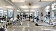 This fitness center at 5350 Burnet Rd in Austin, Texas, offers a spacious, single-floor layout designed to accommodate a full range of workout needs. The open floor plan provides ample room for movement between stations, with a generous footprint that allows for a well-organized arrangement of both cardio and strength-training equipment. The flooring features a contemporary patterned carpet in grey and yellow tones, adding a modern aesthetic throughout the space. Large floor-to-ceiling windows along one wall flood the interior with abundant natural light and offer views of an outdoor area, creating an airy and energizing atmosphere. Full-length mirrors along the opposite wall visually expand the space and allow users to monitor their form during workouts. The ceiling is finished with recessed and pendant lighting, complemented by a ceiling fan for additional air circulation. Equipment includes treadmills, ellipticals, rowing machines, stationary bikes, cable machines, free weights, dumbbells, medicine balls, and various strength machines. A decorative column with a spiral design serves as a visual centerpiece. Colorful circular artwork adorns one wall, contributing to the facility's vibrant character. Glass-paneled doors connect to an adjacent common area, enhancing the sense of openness and accessibility throughout.