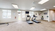 This fitness center on Chapman Dr in Colorado Springs, Colorado, offers a generously proportioned, single-floor space designed to support a well-rounded workout routine. The interior is finished with neutral-toned carpet tiles and light gray walls, creating a clean, modern atmosphere throughout.

Large flush-mounted panel lights line the ceiling, providing bright, even illumination across the entire floor. Multiple windows along the perimeter walls allow natural light to filter in while offering views of the surrounding outdoor greenery, contributing to an open and inviting environment.

The main workout area is equipped with two treadmills, an elliptical trainer, a rowing machine (RowERG), and additional cardio equipment. A wall-mounted flat-screen television is positioned for easy viewing during exercise. On one wall, a mounted pull-up bar and a speed bag station provide functional strength and boxing training options.

Additional features include a dedicated stretch and floor exercise area, a bench with storage, wall-mounted workout reference charts, and clearly posted fitness center rules near the entrance. Interior doors provide access to secondary rooms, likely housing restrooms or storage. The space is also equipped with a fire extinguisher and a clearly marked emergency exit, ensuring safety throughout.