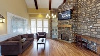 Located at 900 Beverly Hills Rd in Hattiesburg, Mississippi, this single-story space makes a strong architectural impression from the moment you enter. The main living area is anchored by a dramatic floor-to-ceiling stone fireplace, complete with a rustic wood mantle and a mounted flat-screen display above — a striking focal point that draws the eye immediately. Soaring vaulted ceilings with exposed dark wood beams lend a sense of grandeur and openness to the space, while warm hardwood flooring runs throughout, adding both character and continuity. Natural light pours in through a series of white-framed windows dressed with neutral roller shades, creating a bright and welcoming atmosphere. A tasteful chandelier with candle-style lights hangs at the center of the room, complementing the warm tones of the walls and woodwork. Built-in white shelving beside the fireplace offers display and storage space, adding a functional yet decorative touch. The seating area is thoughtfully arranged with comfortable upholstered furniture, making the space feel both inviting and practical. Overall, the design blends rustic charm with clean, classic elements to create a warm and well-appointed environment.