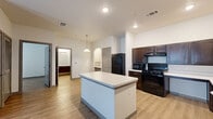 This single-floor, one-bedroom, one-bathroom residence offers a thoughtfully designed layout that maximizes functionality and comfort. Light wood-style flooring flows consistently throughout every room, creating a cohesive and clean aesthetic.

The kitchen serves as a central highlight of the home, featuring rich dark-stained cabinetry, white quartz-style countertops, and a full suite of black appliances including a gas range, microwave, refrigerator, and dishwasher. A freestanding center island provides additional prep space and casual dining potential, while a pendant light above adds a stylish accent. An arched built-in niche offers a charming architectural detail.

The living room is bright and airy, benefiting from natural light through two windows and a glass-paneled exterior door. A ceiling fan overhead ensures year-round comfort. The bedroom is a well-sized retreat, also featuring a ceiling fan, a large window for natural light, and access to a dedicated closet.

The bathroom is designed with accessibility in mind, equipped with grab bars at the tub and toilet, a wall-mounted vanity with a large mirror, and a medicine cabinet with dark wood cabinetry. A utility room rounds out the layout, providing practical storage and laundry accommodations. The space presents a clean, modern, and highly functional living environment.