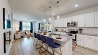 This thoughtfully designed single-floor residence offers a seamless open-concept layout that connects the kitchen, dining, and living areas into one cohesive, light-filled space. The kitchen is a standout feature, showcasing crisp white shaker cabinetry, gray quartz countertops, a classic white subway tile backsplash, and a full suite of stainless steel appliances. A spacious central island with pendant lighting overhead provides ample seating for casual dining and entertaining. Light wood-tone flooring extends throughout the main living areas, enhancing the bright, airy atmosphere. The living room is generously sized with large windows dressed in navy curtains that allow natural light to filter in while offering views to the outdoors. A ceiling fan adds practical comfort to the space. The bedroom is a comfortable retreat featuring plush carpeting, natural light from well-positioned windows, and direct access to a private en-suite bathroom. The bathroom is well-equipped with a dual-sink vanity, gray quartz countertops, white cabinetry, a walk-in glass shower, and wood-tone flooring that ties back to the rest of the home. Throughout the residence, a neutral color palette accented with navy and warm earth tones creates a cohesive, contemporary aesthetic.