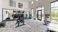 Located at 1876 Star Anise Cir in Apopka, FL, this single-floor fitness center offers a generously sized, thoughtfully designed workout facility. The space is divided into distinct training zones, each catering to a different aspect of physical fitness. Soaring double-height ceilings in key areas lend an open, airy feel, while large floor-to-ceiling windows and glass doors flood the interior with abundant natural light and provide views of the surrounding neighborhood.

The cardio zone is well-appointed with treadmills, ellipticals, stair climbers, and stationary bikes, all positioned to take advantage of the natural light pouring in from surrounding windows. A dedicated strength training area houses a comprehensive dumbbell rack, adjustable benches, and multi-function cable machines, complemented by large wall mirrors that enhance the sense of space. The functional training section features TRX suspension systems, kettlebells, and open floor space ideal for dynamic workouts.

Throughout the facility, a stylish geometric-patterned carpet anchors the clean, neutral color palette of white and light gray walls. Recessed and linear lighting fixtures provide consistent, well-distributed illumination. Motivational artwork adds energy and personality to the space. An additional room off the main floor appears to serve as a private or recovery room, further enhancing the facility's versatility.