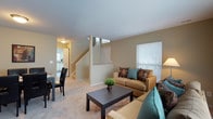 This two-story residence offers a thoughtfully designed layout that maximizes comfort and functionality across both levels. The main floor centers around an open-concept living area bathed in natural light from large windows dressed with classic blinds. The living room provides a warm and inviting atmosphere, featuring neutral-toned carpeting and soft beige walls that create a calm, cohesive aesthetic throughout. A comfortable seating arrangement anchors the space, complemented by a dark wood coffee table that adds a grounding contrast to the light surroundings. A centrally positioned staircase connects the main floor to the upper level, serving as a natural focal point and dividing the living space from the adjoining areas. Adjacent to the living room, a dining area accommodates a table with dark chairs, positioned conveniently near the kitchen. The kitchen is visible in the background, featuring light cabinetry and ample counter space, suggesting a practical and well-appointed cooking environment. The overall ambiance of the main floor is bright, open, and cohesive, with consistent flooring, neutral tones, and clean lines contributing to a sense of spaciousness and ease throughout the home.