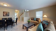 This two-story residence offers a thoughtfully designed layout that maximizes comfort and functionality across both levels. The main floor centers around an open-concept living area bathed in natural light from large windows dressed with classic blinds. The living room provides a warm and inviting atmosphere, featuring neutral-toned carpeting and soft beige walls that create a calm, cohesive aesthetic throughout. A comfortable seating arrangement anchors the space, complemented by a dark wood coffee table that adds a grounding contrast to the light surroundings. A centrally positioned staircase connects the main floor to the upper level, serving as a natural focal point and dividing the living space from the adjoining areas. Adjacent to the living room, a dining area accommodates a table with dark chairs, positioned conveniently near the kitchen. The kitchen is visible in the background, featuring light cabinetry and ample counter space, suggesting a practical and well-appointed cooking environment. The overall ambiance of the main floor is bright, open, and cohesive, with consistent flooring, neutral tones, and clean lines contributing to a sense of spaciousness and ease throughout the home.