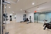Located at 32 SE Second Ave in Delray Beach, FL, this well-appointed fitness center offers a comprehensive workout environment across a single, open floor. The space is designed with a clean, modern aesthetic, featuring crisp white walls, light wood-style flooring, and recessed lighting complemented by ceiling fans that maintain a comfortable atmosphere throughout.

The main workout area is generously sized, accommodating a diverse selection of equipment including a multi-station cable machine, weight benches, a rowing machine, stationary and spin bikes, and a dedicated rack of dumbbells. Full-length mirrors line portions of the walls, enhancing the sense of openness and allowing users to monitor their form. Mounted flat-screen televisions provide entertainment throughout the space.

A separate, glass-enclosed room houses additional cardio equipment, including spin bikes, offering a more private workout setting. A dedicated alcove features treadmills and an elliptical trainer alongside a floor-to-ceiling mirror. Decorative artwork adds a touch of personality to the otherwise streamlined interior. Lockers and utility panels are neatly integrated along one wall. The overall ambiance is bright, functional, and professionally outfitted, making this fitness center a well-rounded facility for a full range of exercise needs.