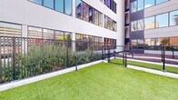 Located at 555 E Main St in downtown Stamford, Connecticut, this outdoor dog run is a thoughtfully designed amenity space set against the backdrop of a modern multi-story residential building. The space features a generously sized, elongated stretch of vibrant artificial turf that provides a clean, low-maintenance surface for pets to roam and play year-round. The run is fully enclosed by a sturdy black metal fence with secure gated entry points, ensuring the safety of all animals within. Raised planting beds line portions of the perimeter, adding greenery and visual appeal with shrubs and ornamental grasses that soften the urban setting. Practical amenities are integrated throughout, including a pet water station and waste disposal stations equipped with bags and receptacles, making the space fully functional for daily use. Young trees are interspersed along the length of the run, offering shade and a natural atmosphere. The surrounding cityscape — including neighboring brick buildings and open sky — gives the space an open, airy feel despite its urban location. The dog run is divided into sections by interior fencing with gated access, allowing for flexible use. Overall, this space offers a well-maintained, accessible, and inviting outdoor environment for pets and their owners in the heart of downtown Stamford.