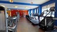 This single-floor fitness center offers a comprehensive workout environment designed to accommodate a range of exercise routines. The space is visually striking, featuring bold blue accent walls paired with a vivid red wall at the far end, creating an energetic and motivating atmosphere. The flooring consists of durable hardwood-style material, providing a clean and practical surface throughout.

Along one side of the room, a row of modern treadmills and elliptical machines is neatly arranged, each equipped with individual digital display screens. Opposite these cardio stations, additional cardio equipment lines the other side, maximizing the use of the space. Natural light filters in through a series of windows along the right-hand wall, brightening the interior and contributing to an open, airy feel.

Toward the back of the room, a dedicated strength training area features cable machines, a multi-functional rack, and various resistance equipment, offering users a full range of workout options. Overhead, pendant-style and flush-mount light fixtures provide consistent illumination throughout the space. The layout is organized and spacious, allowing for comfortable movement between equipment stations.