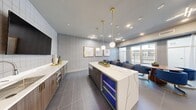 Located at 3240 W Harmon Ave in Las Vegas, NV, this single-floor multi-purpose lounge and entertainment space offers a sophisticated, contemporary interior designed for both relaxation and social gatherings. The layout seamlessly integrates several distinct functional zones across a generous open floor plan. A sleek wet bar anchors one side of the space, featuring a striking white marble-veined island with illuminated wine coolers, warm wood cabinetry with gold hardware, and a wall-mounted flat-screen TV. Pendant globe lights with brushed gold fixtures hang above, adding an elegant, modern touch. Adjacent to the bar, a formal dining area accommodates a round wood table surrounded by plush royal blue velvet chairs, framed by large floor-to-ceiling windows and glass doors that flood the space with natural light and provide access to an outdoor balcony. A lounge area is furnished with coordinating blue striped accent chairs and a gold drum coffee table atop a textured area rug, with decorative mirrors and framed artwork adorning the soft gray chevron-patterned wallcoverings. A dedicated media lounge features built-in shelving, a large wall-mounted TV, and a marble-finished credenza. Large-format gray tile flooring unifies the entire space, while recessed and geometric ceiling light fixtures provide a refined, polished ambiance throughout.