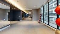 Located at 65 Broadway Ave in Toronto, this well-appointed fitness facility offers a generously sized, single-floor layout designed to support a full range of exercise activities. The space is divided into two distinct zones: a comprehensively equipped gym area and an open, flexible studio space.

The main gym area is an expansive, elongated room outfitted with a wide selection of cardio machines, including multiple treadmills, as well as strength training equipment, cable machines, free weights, and adjustable benches. Full-length mirrors line the walls, amplifying the sense of space and allowing users to monitor their form. A warm wood-paneled ceiling with recessed lighting adds a refined, modern aesthetic, while large floor-to-ceiling windows along the exterior wall flood the space with natural light and provide views of the surrounding Toronto cityscape.

The secondary studio area is an open, multi-purpose room finished with the same durable rubberized tile flooring. It features glass partitioning that visually connects it to the main gym, along with additional floor-to-ceiling windows that overlook an outdoor terrace. This versatile space is well-suited for stretching, group fitness, yoga, or functional training. The overall design balances a sleek, contemporary aesthetic with practical, high-performance functionality throughout.