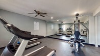This fitness center is designed to support a well-rounded exercise routine within a clean, functional layout. The space is finished with durable rubber flooring throughout, providing traction and cushioning suitable for a variety of workouts. Gray-painted walls and recessed lighting create a focused, modern atmosphere, while ceiling fans and a wall-mounted HVAC unit ensure comfortable temperature regulation during use.

The cardio area is equipped with multiple treadmills featuring digital display consoles, as well as upright and recumbent stationary bikes, catering to varying fitness preferences and intensity levels. A multi-station cable machine serves as the centerpiece for strength training, complemented by additional resistance equipment and a workout bench. Floor-to-ceiling mirrors line one wall, enhancing the sense of openness while allowing users to monitor their form.

Natural light filters in through a window, and a glass-panel door provides direct exterior access. Posted fitness center rules are clearly displayed near the entrance. Structural support columns are incorporated into the layout without significantly impeding the usable floor space. Overall, the fitness center offers a practical, well-maintained environment equipped to meet a broad range of exercise needs.