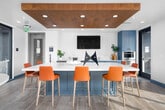 This contemporary amenity lounge is designed to impress, offering an open-plan layout that seamlessly connects a fully equipped kitchen, a dining area, a lounge, and a generous outdoor terrace. The kitchen is appointed with striking teal cabinetry, white quartz countertops, a marble-inspired backsplash, hexagonal tile flooring, and stainless steel appliances, including a wall oven and beverage cooler. A large white island with bar-height seating in bold orange serves as a central gathering point, accented by a warm wood-paneled ceiling overhead that adds a refined, natural touch. The open lounge area features a plush sectional sofa in neutral tones, layered with blue and teal accent pillows atop a patterned area rug, complemented by navy blue accent chairs and a sleek glass coffee table. Floor-to-ceiling black-framed sliding glass doors flood the interior with abundant natural light and provide direct access to the expansive rooftop terrace. The terrace is furnished with outdoor dining tables, bar-height seating, and lounge chairs, all framed by sweeping skyline views. Throughout the space, the design palette of blues, oranges, and warm wood tones creates a cohesive, energetic, and inviting atmosphere ideal for gathering and entertaining.