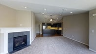This single-floor residence showcases an open-concept layout that seamlessly connects the living area and kitchen, creating a spacious, cohesive environment. The living room is anchored by a classic white-mantled fireplace with dark tile surround, providing a focal point that complements the neutral beige walls and plush carpeted flooring. Vaulted ceilings rise above the central living and kitchen space, enhancing the sense of openness, while a ceiling fan ensures comfortable airflow throughout.

The kitchen is outfitted with dark espresso cabinetry that contrasts elegantly against a light tile backsplash. A central island with a dark finish offers additional prep and seating space, while pendant lighting overhead adds a stylish, functional touch. Recessed lighting is distributed throughout both the living and kitchen areas, providing consistent, well-balanced illumination. Stainless steel appliances complete the modern aesthetic of the kitchen. Dark hardwood flooring in the kitchen transitions smoothly into the carpeted living area, delineating the two spaces while maintaining visual harmony across the open floor plan.