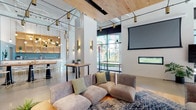 Located at 500 W Broad St in Columbus, Ohio, this single-floor commercial space delivers a contemporary and welcoming atmosphere through a thoughtfully designed open-plan layout. The interior is anchored by warm natural wood ceiling panels that contrast beautifully with crisp white walls and polished concrete flooring, creating a refined yet approachable aesthetic.

The lounge area is a central highlight, featuring plush modular sofas in soft earth tones, layered over a patterned area rug, and accented with sage green throw pillows. A large wall-mounted projection screen and a sleek linear gas fireplace provide both entertainment and comfort to the space. Nearby, tall bistro tables offer casual seating options for smaller gatherings.

Natural light filters in through generous floor-to-ceiling windows, which also frame a vibrant, colorful mural that enlivens the interior with artistic character. The bar area, visible in the background, is outfitted with pendant lighting, bar-height seating, and a well-stocked display, making it an inviting social hub.

Layered lighting throughout—including track lighting, cylindrical pendant fixtures, and warm wall sconces—ensures a dynamic ambiance suitable for a variety of occasions. The space combines functional design with curated décor to create a polished, communal environment ideal for socializing and events.