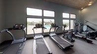 Located on Calle Cancuna in Carlsbad, California, this single-story fitness center is designed to support a comprehensive range of workout routines. The space is divided into distinct functional zones, each thoughtfully outfitted with professional-grade equipment.

The cardio area features multiple treadmills, elliptical trainers, and a stair climber positioned alongside a generous bank of large windows. These windows frame views of an outdoor pool area and the surrounding landscape, providing natural light and an open, airy atmosphere. A warm wood-paneled ceiling adds a distinctive architectural detail to this section of the facility.

The strength training area is spacious and well-lit with recessed lighting, accommodating a variety of cable machines, a multi-functional rack with suspension trainers, free weights, and an adjustable bench. Teal accent walls contrast with neutral gray tones throughout, contributing a modern, energetic aesthetic. Durable rubber flooring covers the entire space, offering both safety and functionality.

A mirrored wall in the main strength zone visually expands the room while allowing users to monitor their form. Windows along one wall invite additional natural light and outdoor views. The overall layout is organized and open, providing ample room to move freely between equipment stations.