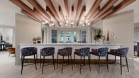 This single-floor amenity space is designed with a refined, contemporary aesthetic that blends warm natural materials with bold decorative accents. The centerpiece of the lounge area is a striking ceiling treatment featuring exposed wood beams arranged in a geometric pattern, complemented by a brass-accented sputnik chandelier that casts ambient light across the room. A white quartz bar counter lined with upholstered navy patterned barstools serves as a social hub, with bay windows behind it framing views of the lush outdoor greenery.

Light wood-look flooring runs throughout, unifying the open layout. A fully equipped kitchenette anchors one side of the space, showcasing a dramatic gold mosaic tile backsplash, wood-toned lower cabinetry, crisp white upper cabinets, floating shelves, a stainless steel dishwasher, and a wine refrigerator. A textured white stone accent wall adds organic contrast in the dining nook, where round bistro-height tables and mixed seating create an intimate gathering area illuminated by wall sconces. Dark-framed glass doors connect to an adjacent corridor, while a built-in leather banquette and wood dining table provide additional seating, rounding out this elegantly layered communal environment.