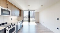 Located in the heart of Midtown Atlanta at 1382 Peachtree St NE, this modern studio offers a thoughtfully designed open-concept layout combining the kitchen and living area into one cohesive space. The unit is anchored by dramatic floor-to-ceiling windows framed in sleek black, flooding the interior with abundant natural light and delivering sweeping views of the Atlanta skyline and surrounding cityscape. Light-toned hardwood-style flooring runs throughout, complementing the clean white walls and contemporary aesthetic.

The kitchen is well-appointed with a full suite of stainless steel appliances, including a refrigerator, gas range, built-in microwave, and dishwasher. Two-toned cabinetry — warm wood upper cabinets paired with matte slate-blue lower cabinets — adds visual depth, while a white tile backsplash and light countertops complete the modern look. A stacked in-unit washer and dryer are conveniently tucked behind a dedicated closet door.

The bathroom features large-format beige tile flooring, a glass-enclosed walk-in shower with full-height dark tile surround, a wall-mounted illuminated mirror, and a floating wood vanity with an undermount sink. A ceiling fan in the living area and recessed lighting throughout round out this polished, functional space.