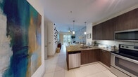 This thoughtfully designed single-floor residence offers a seamless open-concept layout that connects the kitchen, dining, and living areas into one cohesive and functional space. The kitchen is equipped with dark wood cabinetry, stainless steel appliances, a subway tile backsplash, and a granite-topped peninsula with bar seating — ideal for casual dining or entertaining. Pendant lighting and track fixtures add warmth and dimension throughout.

The living area is anchored by a striking geometric accent wall in soft grey and white, which draws the eye toward the sliding glass doors that open onto a private balcony with outdoor views. Light-toned tile flooring runs throughout the main living spaces, enhancing the sense of openness and brightness.

The bedroom continues the geometric design theme with a vibrant teal and grey accent wall, creating a contemporary and energetic atmosphere. Carpeted flooring adds comfort underfoot, and a window provides natural light and outdoor views.

The bathroom features large-format tile, a full soaking tub with shower, a dark wood vanity with granite countertop, and a soft blue accent wall that adds a calming touch. The entryway is welcoming and well-appointed, featuring decorative wall art and direct access to a louvered closet. Throughout the space, a cohesive modern aesthetic is maintained with curated decor and design elements.
