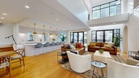 Located at 2001 Dabney Rd in Richmond, VA, this thoughtfully designed two-floor space offers a generous layout spread across two well-appointed levels. The ground floor makes an immediate impression with its soaring double-height ceilings, expansive floor-to-ceiling black-framed glass doors, and warm hardwood flooring that flows seamlessly throughout. The open-concept kitchen and living area serves as the heart of the space, anchored by a sweeping curved white quartz island with fluted detailing and seating for multiple guests, illuminated by a striking row of brass pendant lights. White cabinetry, arched mirror accents, and a matte black sink give the kitchen a refined yet approachable character. The adjacent living areas offer distinct atmospheres — one bathed in natural light from large exterior-facing windows with views of the surrounding development, and another featuring a dramatic white-painted brick accent wall with a large mounted display screen and soaring ceilings. The upper floor provides additional lounge space with bold design choices, including deep teal board-and-batten walls, globe sconces, and curated botanical artwork. A connecting hallway on the upper level overlooks the ground floor below, reinforcing the open, airy feel of the entire space. Rich textures, layered lighting, and cohesive modern design define the overall ambiance.