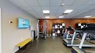 This fitness facility offers a thoughtfully designed workout environment that balances functionality with a welcoming ambiance. The space features a drop-panel ceiling with recessed fluorescent lighting that ensures bright, even illumination throughout the room. The flooring consists of durable, speckled rubber gym matting, providing both safety and comfort during exercise.

The strength training area is anchored by a multi-function cable machine with an adjustable bench, ideal for a wide range of resistance exercises. A wall-mounted flat-screen television is positioned prominently for entertainment or instructional viewing during workouts. A simple wooden bench nearby offers a convenient spot for stretching or rest.

The cardio section features treadmills positioned along one side of the room, allowing users to exercise while facing the open space. A bold terracotta-orange accent wall adds warmth and energy to the environment, complemented by decorative touches including potted orchids, tall grasses, and a modular shelving unit with organized storage cubbies. A floor lamp adds a softer lighting element, contributing to a more inviting atmosphere.

Overall, this fitness space is well-suited for a range of workout routines, combining essential equipment with a clean, organized layout and a warm, motivating aesthetic.
