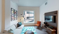 This 1-bedroom, 1-bathroom residence offers a thoughtfully designed single-floor layout that balances comfort and modern style. The living area serves as the heart of the space, featuring clean white walls and smooth ceilings that create an open, airy atmosphere. A large picture window allows natural light to fill the room while framing views of the surrounding urban environment. Light wood-style flooring runs throughout, lending warmth and continuity to the space. The living room is arranged for both relaxation and functionality, centered around a plush sectional sofa accented with decorative throw pillows in teal and navy tones. A vibrant teal chevron-patterned area rug anchors the seating area, complemented by a retro-inspired orange accent chair that adds a pop of color. A sleek wall-mounted media console with a flat-screen television provides an organized entertainment focal point. A distinctive multi-arm floor lamp adds both character and ambient lighting. A curated gallery wall of framed graphic art prints brings personality and visual interest to the room. The overall ambiance is contemporary and inviting, combining neutral base tones with bold accent colors for a space that feels both stylish and livable.