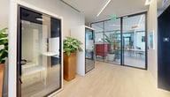 Located at 51 NW 28th St in Miami's vibrant Wynwood neighborhood, this contemporary commercial space offers a thoughtfully designed single-floor layout that balances open-plan functionality with private work zones. Light hardwood flooring runs throughout, complementing the clean, modern aesthetic defined by floor-to-ceiling windows and sheer white curtains that flood the interior with natural light and frame views of lush palm trees and outdoor greenery beyond.

The space features sleek glass-walled partitions and private acoustic phone or focus pods — compact, self-contained booths equipped with built-in desks and soundproofing — ideal for focused individual work or confidential calls. An open lounge-style area with vibrant red seating and abundant indoor plants adds warmth and color, while the corridor leading deeper into the space is adorned with a bold, colorful abstract artwork, adding a distinctly creative, gallery-like quality consistent with the surrounding Wynwood arts district.

Elevator access is available, with hallways finished in recessed linear lighting that enhances the professional atmosphere. A water station and safety fixtures are conveniently integrated into the design. The interplay of natural light, curated art, greenery, and modern architectural elements creates a dynamic, inspiring environment well-suited for a coworking, creative office, or professional services use.