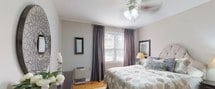 This thoughtfully designed single-floor residence showcases a cohesive aesthetic centered on a soft, neutral palette of warm grays and creamy whites throughout. Warm hardwood flooring flows seamlessly across each room, providing an inviting and unified foundation.

The living room serves as the heart of the home, bathed in natural light from multiple windows dressed with sheer, floor-length curtains. A tufted charcoal sofa anchors the space, complemented by a pair of accent chairs, a large upholstered ottoman, and a striking circular wood-framed mirror. Decorative artwork and elegant table lamps complete the refined atmosphere.

The primary bedroom is a serene retreat featuring large windows with layered gray drapes that filter natural light beautifully. A generously sized bed with a tufted upholstered headboard takes center stage, flanked by matching nightstands and lamps, while a ceiling fan overhead adds comfort. Hardwood floors extend beneath a classic lattice-patterned area rug.

A versatile secondary room offers flexibility, currently styled as a relaxing sitting or guest space with a daybed, patterned area rug, and decorative wall accent, with natural light provided by a draped window.

The intimate dining nook features a round wood table with zebra-print accent chairs, a framed wall mirror, and a window that lets in ample natural light — ideal for casual dining.