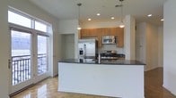 This single-floor residence is centered around an open-concept kitchen that serves as the focal point of the living area. The kitchen is outfitted with a full suite of stainless steel appliances, including a French door refrigerator with an ice and water dispenser, a gas range, and an over-the-range microwave. Warm-toned wood cabinetry provides ample upper storage, while the expansive peninsula counter features a dark granite surface, offering generous prep space and a natural division between the kitchen and the adjoining living area. Pendant lighting hangs above the peninsula, complementing the recessed ceiling lights to create a well-lit environment. The flooring throughout features a classic parquet wood pattern, adding texture and warmth to the interior. A large window and a glass-paneled door lead to a private balcony, allowing natural light to fill the space and offering views of the exterior surroundings. A walk-in closet is visible adjacent to the kitchen, providing additional storage. White walls and clean trim contribute to a bright, neutral aesthetic throughout.