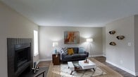 This single-story space presents a well-appointed living area that combines comfort and contemporary style. The living room is characterized by warm wood-style flooring that anchors the space, complemented by neutral greige walls that create a clean and versatile backdrop. A textured popcorn ceiling adds a classic architectural detail overhead.

The room is thoughtfully arranged around a dark charcoal sofa accented with decorative pillows in chevron, mustard yellow, and teal tones, adding pops of color to the neutral palette. A modern white coffee table with mid-century legs serves as the focal point of the seating area, paired with a patterned area rug that defines the space.

Natural light filters in through a window fitted with horizontal blinds on the left side of the room, while two stylish floor and table lamps provide warm ambient lighting. A framed abstract artwork in warm earth tones hangs above the sofa, tying the color scheme together cohesively. Dark wood side tables and baseboard trim provide rich contrast against the lighter walls. The overall ambiance is relaxed yet polished, offering a functional and inviting living environment.