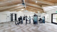 Located at 1440 S Val Vista Dr in Mesa, Arizona, this fitness center is a thoughtfully designed, single-floor facility offering a generous workout space for residents. The room's most striking architectural feature is its soaring vaulted ceiling, detailed with natural wood tongue-and-groove paneling and exposed wooden beams, lending a warm, lodge-inspired aesthetic that sets it apart from a typical gym environment. Recessed lighting throughout ensures the space is well-lit, complementing the abundant natural light that streams in through multiple large windows.

The flooring features a stylish herringbone-patterned carpet tile in neutral tones, adding texture and visual interest underfoot. The equipment layout is spacious and organized, encompassing a comprehensive selection of strength-training machines, a cable crossover system, free weights with a dumbbell rack, medicine balls, TRX suspension trainers, a rowing machine, and a dedicated stretching area with wall-mounted mirrors.

On the cardio side, treadmills, stair climbers, and elliptical machines line a windowed wall that looks directly out onto an outdoor pool and courtyard area, providing a refreshing view during workouts. Framed artwork on the crisp white walls and a ceiling fan add finishing touches that enhance the overall ambiance, making this fitness center both functional and inviting.
