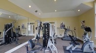 Located at 725 Heather Park Dr in Garner, NC, this dedicated fitness center offers a well-equipped, single-floor space designed to support a well-rounded exercise routine. The room features a tray ceiling with recessed lighting that keeps the space bright and energized, while warm yellow walls contribute to an inviting, motivating atmosphere. Large windows dressed with blinds allow natural light to filter in, enhancing the overall openness of the room. A full-length wall mirror spans one side, creating a sense of expanded space and allowing users to monitor their form during workouts. The facility is outfitted with a multi-station cable strength machine, ideal for a variety of resistance training exercises, alongside cardio equipment including a treadmill and an elliptical or stationary bike. Motivational fitness posters are displayed on the walls, adding an energetic touch to the environment. The carpeted flooring with rubber mat sections provides both comfort and safety underfoot. The room is accessible via French-style glass panel doors, adding a polished architectural detail to the functional space. Overall, this fitness center is a compact yet comprehensive amenity, thoughtfully designed to meet everyday health and wellness needs.
