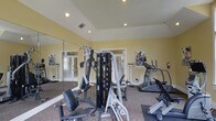 Located at 725 Heather Park Dr in Garner, NC, this dedicated fitness center offers a well-equipped, single-floor space designed to support a well-rounded exercise routine. The room features a tray ceiling with recessed lighting that keeps the space bright and energized, while warm yellow walls contribute to an inviting, motivating atmosphere. Large windows dressed with blinds allow natural light to filter in, enhancing the overall openness of the room. A full-length wall mirror spans one side, creating a sense of expanded space and allowing users to monitor their form during workouts. The facility is outfitted with a multi-station cable strength machine, ideal for a variety of resistance training exercises, alongside cardio equipment including a treadmill and an elliptical or stationary bike. Motivational fitness posters are displayed on the walls, adding an energetic touch to the environment. The carpeted flooring with rubber mat sections provides both comfort and safety underfoot. The room is accessible via French-style glass panel doors, adding a polished architectural detail to the functional space. Overall, this fitness center is a compact yet comprehensive amenity, thoughtfully designed to meet everyday health and wellness needs.