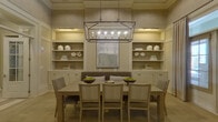 This thoughtfully designed single-floor space exudes sophistication through its cohesive neutral palette, textured grasscloth-style wallcoverings, and wide-plank hardwood flooring that flows throughout. The welcoming foyer sets an elegant tone with soaring ceilings, a statement lantern-style chandelier, a central round accent table adorned with fresh orchids, and flanking French doors with glass panes that invite natural light deep into the interior. Upholstered wing chairs and decorative ceramic garden stools complete the refined entryway. The formal dining area is a standout feature, anchored by a generously sized light wood dining table surrounded by upholstered striped chairs and a tufted bench. A dramatic rectangular iron-and-glass chandelier hangs overhead, casting warm ambient light. Flanking the dining space, symmetrical built-in shelving units with cabinetry below display curated ceramic accents and artwork, adding depth and visual interest. A built-in workstation is seamlessly integrated into one wall, featuring open shelving, a compact desk surface, and a comfortable upholstered chair — offering a functional yet stylish workspace. The overall ambiance is calm and cohesive, balancing classic architectural detailing with contemporary furnishings for a polished, livable aesthetic.