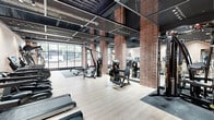 This fitness facility occupies a single open floor and is designed with a sharp industrial aesthetic that combines raw materials with modern finishes. Exposed brick columns rise throughout the space, contrasting against light wood-look flooring and a dark perforated metal ceiling that reveals structural elements and visible ductwork above. Large floor-to-ceiling windows line the street-facing wall, flooding the interior with abundant natural light and offering views of the surrounding urban streetscape.

The workout floor is thoughtfully arranged to accommodate a diverse range of training modalities. True-brand treadmills and a stair climber are positioned along one end of the space, while cable machines, multi-functional strength stations, adjustable benches, and a dumbbell rack with resistance bands occupy the central and surrounding areas. Rubber mat zones demarcate dedicated strength and free-weight training sections, contrasting with the light flooring elsewhere.

A bold orange accent wall bearing the phrase "Hard Hittin'" serves as a striking focal point, reinforcing the facility's energetic brand identity. Full-length mirrors are integrated along portions of the walls, enhancing the sense of openness. Recessed and track lighting supplements the natural light, ensuring a well-lit environment throughout. The overall ambiance is motivating, modern, and purposefully designed for performance-focused training.