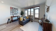 This single-floor unit showcases a thoughtfully designed open-concept layout with consistent light wood-tone flooring running throughout. The living area is a bright, welcoming space anchored by a bank of large windows with warm wood-trimmed frames, offering expansive urban views and abundant natural light. A chevron-patterned area rug and a balcony visible through the windows add texture and depth to the room.

The kitchen flows seamlessly from the living area, featuring a crisp two-tone design with white upper cabinets and dark navy lower cabinets. Stainless steel appliances, including a full-size refrigerator, electric range, microwave, and dishwasher, provide a fully equipped cooking space. Recessed lighting illuminates the area effectively.

The bedroom is a calm, neutral retreat with a sliding dark barn-style door leading to a built-in closet accented by warm wood trim, maintaining the unit's consistent design language.

The bathroom is clean and functional, finished in white with a grey quartz-topped vanity, undermount sink, modern cabinetry, and light wood-look flooring. A curtained shower and framed wall art contribute to its polished, contemporary feel.