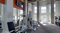 This fitness center is designed to provide an energizing and functional workout environment. The space is characterized by impressively tall ceilings that lend a sense of openness and grandeur, enhanced by large, floor-to-ceiling windows that flood the room with natural light and offer views of the outdoor surroundings. The architecture features clean white walls accented by bold orange detailing, giving the space a modern and motivating aesthetic.

The gym is well-appointed with a variety of strength training cable machines and resistance equipment, thoughtfully arranged to allow ample room for movement and exercise. Large mirrors line one wall, visually expanding the space while allowing users to monitor their form during workouts. A wall-mounted flat-screen television adds a convenient entertainment option during training sessions.

The flooring features a rich, dark hardwood finish that contrasts with the bright walls, contributing to an upscale, polished look. The combination of high ceilings, natural light streaming through the oversized windows, and carefully selected equipment makes this fitness center both practical and aesthetically impressive.