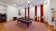 Located at 2110 Pickens St. in Columbia, SC, this single-floor amenity lounge is designed for recreation and socializing. The space is anchored by a full-size pool table with ornate wooden legs, set atop a warm-toned area rug that contrasts beautifully with the rich hardwood flooring throughout. Bold red drapes frame large windows, allowing natural light to fill the room while adding a striking pop of color against the clean white walls.

A built-in wall niche serves as a decorative bookshelf, adding a charming, residential touch. High-top tables with bar-height seating are positioned near the windows, creating ideal spots for casual conversation. Stylish globe pendant fixtures and recessed ceiling lights provide layered illumination throughout.

Entertainment options abound, including a classic upright multi-game arcade cabinet, a tabletop arcade unit, and a wall-mounted flat-screen TV paired with a cue rack for pool players. A seating nook with two soft teal lounge chairs and a round pedestal table offers a relaxed retreat.

A striking floor-to-ceiling black-framed glass partition wall separates an adjacent corridor, adding an industrial-chic architectural element while maintaining a sense of openness and visual connectivity throughout the space.