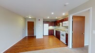This single-floor unit offers a practical and well-appointed layout designed for comfortable everyday living. The open-concept kitchen and living area serves as the heart of the space, seamlessly connecting functional cooking and relaxing zones. The kitchen is equipped with rich, dark cherry wood cabinetry that provides generous storage, complemented by white appliances including a refrigerator, electric range, built-in microwave, and a double-basin sink beneath recessed lighting. The countertops are light-toned, offering a clean contrast to the warm cabinetry. Warm hardwood flooring extends throughout the main living area, adding a sense of richness and continuity, while transitioning to carpet in adjoining areas. Recessed ceiling lights and flush-mount fixtures provide ample illumination throughout. The entryway features a solid wood front door, creating a welcoming and secure entry point. The unit also includes additional doorways leading to separate rooms or utility spaces, contributing to a well-organized and efficient floor plan. The neutral wall tones and clean finishes throughout give the space a bright, move-in-ready ambiance suited to a range of furnishing styles.