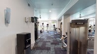 This fitness center is a comprehensively outfitted facility designed to support a wide range of workout routines. The space is organized into distinct functional zones across a single floor, all unified by a stylish patterned carpet in deep blues and grays that adds visual texture throughout. Crown molding and recessed lighting lend a polished, finished look to the interior.

The cardio section lines the perimeter near large windows that welcome abundant natural light and offer views of surrounding greenery, creating an energizing atmosphere. Equipment includes elliptical trainers, upright and recumbent stationary bikes, and treadmills, each outfitted with individual monitor screens. A strength training area features Precor cable machines, selectorized resistance machines, and seated weight benches, complemented by floor-to-ceiling mirrors that enhance the sense of space and allow users to monitor form.

A dedicated free weights section provides a full rack of dumbbells, while the functional fitness zone stocks stability balls in multiple sizes, medicine balls, and rolled exercise mats on organized storage racks. Amenity stations along the walls include a water dispenser, paper towel and sanitizing wipe dispensers, and a basket for towels. A posted rules board highlights gym policies, including equipment care and safety guidelines, reflecting a well-maintained and community-oriented facility.