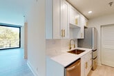 This single-floor space showcases a clean, contemporary design characterized by an all-white palette, light wood-look tile flooring, and abundant natural light. The open living area centers around a large sliding glass door that leads to a balcony with views of mature trees, creating a seamless connection between interior and exterior. A floor-to-ceiling window in one of the bedrooms further reinforces this indoor-outdoor relationship, flooding the room with natural light and green canopy views.

The galley-style kitchen is a standout feature, appointed with white shaker cabinetry, quartz countertops, a white subway tile backsplash, and a full suite of stainless steel appliances. Gold hardware throughout — including cabinet pulls, a high-arc bronze kitchen faucet, and a deep dual-basin sink — adds a refined, upscale touch that is carried consistently across the unit.

Multiple bedrooms feature the same light tile flooring and minimalist white walls, each benefiting from generous window placement and tree-lined outdoor views. The bathroom continues the gold-accented theme with a white vanity, round mirror with a gold frame, gold fixtures, and elegant lighting. The entry area offers a practical layout with direct kitchen access. Overall, the space conveys a cohesive, modern aesthetic with thoughtful detailing throughout.