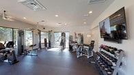 Located at 8100 US 98 in Pensacola, Florida, this fitness center is a well-appointed, single-floor facility designed to support a full range of workout routines. The space is bathed in natural light, courtesy of large windows on multiple walls that offer views of the surrounding outdoor areas, including a nearby pool and landscaped grounds. Recessed lighting and ceiling fans throughout ensure a comfortable and well-illuminated environment at all times of day.

The gym floor is covered in durable rubber flooring, providing a safe and functional surface for all types of exercise. The strength training area is thoroughly equipped with multiple cable and resistance machines, a comprehensive dumbbell rack, and adjustable benches to accommodate a variety of exercises. The cardio section features treadmills, an elliptical trainer, a stationary bike, and a rowing machine, offering diverse options for cardiovascular workouts.

Additional amenities include a wall-mounted flat-screen television, a dedicated storage rack for yoga mats and foam rollers, and a mirror for form monitoring. Glass doors open directly to the outdoor pool area, seamlessly connecting the interior fitness space to the exterior amenities. The clean, white walls and open layout contribute to an energizing and functional atmosphere suited for community fitness use.