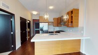 This single-floor residence showcases a thoughtfully designed open-concept layout that centers around a stunning kitchen. The kitchen is a standout feature, boasting warm bamboo-toned wood cabinetry that extends from base to upper wall units, complemented by glass-front display cabinets that add a refined touch. A generous kitchen island with a crisp white countertop and an undermount sink serves as both a functional workspace and a natural gathering point. Overhead, two elegant globe-style pendant lights with candelabra detailing provide warm, ambient illumination, enhancing the inviting atmosphere. Stainless steel appliances, including a French door refrigerator with an ice dispenser and a gas range with a ventilation hood, add a professional-grade quality to the space. A subtle mosaic tile backsplash in cool tones provides a tasteful contrast to the warm wood tones throughout. Rich, dark hardwood floors run throughout the space, grounding the design with a sense of warmth and sophistication. The overall ambiance is modern yet welcoming, with a clean, well-appointed aesthetic that balances practicality with contemporary style.