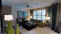 This single-floor space showcases a thoughtfully designed living room that balances comfort with contemporary style. The room is anchored by a large sofa dressed in warm tan upholstery, complemented by woven armchairs adorned with an array of decorative pillows in teal, green, and navy tones. A bold dark-finished coffee table serves as the centerpiece, adding a striking contrast to the light, neutral-toned carpet that spans the floor.

Large windows dressed with layered blinds and teal drape panels flood the room with natural light, offering views of lush greenery outside. Crown molding frames the ceiling, which features a ceiling fan that adds both function and a classic touch. The walls are kept in a soft, warm neutral, providing an ideal backdrop for the two framed abstract artworks in cool blues and greens that anchor the main wall.

Ambient lighting is thoughtfully incorporated through table lamps, creating a warm and inviting atmosphere. Decorative accents—including geometric green vases and yellow floral arrangements—add vibrant pops of color throughout. Overall, the space presents a cohesive, well-curated environment that is both visually engaging and functionally comfortable.