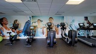 This single-floor fitness space is designed to support cardio-focused workouts in a clean, functional environment. The room is equipped with Life Fitness machines, including two elliptical trainers and a recumbent stationary bike, each featuring individual display screens for tracking workout metrics. The flooring consists of dark rubber gym matting, which provides durability and cushioning underfoot. The ceiling features a standard drop-tile grid with recessed lighting and ventilation panels, ensuring adequate illumination and airflow throughout the space. A prominent full-wall photographic mural depicting active individuals exercising serves as a dynamic visual focal point, adding energy and motivation to the room's atmosphere. The overall design is straightforward and utilitarian, prioritizing functionality over decorative detail. The layout allows for comfortable spacing between machines, making the space accessible and easy to navigate. This fitness room appears well-suited as an amenity within a larger facility, offering residents or guests convenient access to quality cardio equipment in a tidy, purpose-built setting.