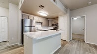 This single-floor residence offers a thoughtfully designed interior with a clean, contemporary aesthetic throughout. The kitchen is a standout feature, showcasing gray cabinetry with brushed hardware, a white subway tile backsplash, gleaming white countertops, and a full suite of stainless steel appliances including a refrigerator, dishwasher, microwave, and range. A peninsula island provides additional prep space and separates the kitchen from the open living area, which is finished with light wood-look plank flooring that extends seamlessly throughout the main living spaces. The living area is bright and open, benefiting from a ceiling fan and a window fitted with vertical blinds that allows natural light to filter in, with access to an outdoor area. The bedroom is well-proportioned, featuring the same wood-look flooring and a ceiling fan. It connects directly to a generously sized walk-in closet outfitted with multi-tier wire shelving and carpeted flooring, offering excellent storage capacity. The bathroom is finished with wood-look tile flooring, a gray vanity with a white countertop, and a full tub-shower combination with white tile surround and brushed nickel fixtures, delivering a polished and functional space.