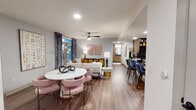This thoughtfully designed single-floor residence combines modern aesthetics with functional living across a well-planned open layout. The living and dining area flow seamlessly together, anchored by warm wood-look flooring, recessed lighting, and a ceiling fan overhead. The dining space features a round white table surrounded by blush pink velvet chairs with gold accents, while the living room offers a neutral sofa, eclectic side tables, and large windows dressed in velvet drapes that invite natural light. The kitchen is a standout feature, showcasing two-tone cabinetry — warm wood uppers paired with dark lower cabinets — complemented by a bold dark tile backsplash, dark granite countertops, stainless steel appliances, and a breakfast bar with seating. The primary bedroom is distinguished by a deep navy accent wall, ceiling fan, hardwood-style flooring, and a framed window with blinds. A second bedroom presents a warm, inviting atmosphere with neutral tones and contemporary artwork. Both bathrooms are finished with dark granite countertops, wood-toned vanities, framed mirrors, and clean white fixtures — one featuring a full vanity with ample counter space, the other a sleek single-sink setup with stylish lighting. Throughout, the design palette blends bold accents with neutral tones for a cohesive, contemporary feel.