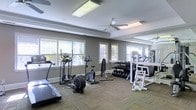 Located at 8160 County Road 64 in Daphne, Alabama, this fitness center offers a well-equipped, single-floor exercise facility designed to support a comprehensive workout experience. The space features a thoughtfully arranged layout with a wide variety of cardio machines, including ellipticals, stationary bikes, and treadmills, alongside a full dumbbell rack and multi-use strength training equipment. The interior is finished with neutral gray walls accented by crisp white crown molding and baseboards, lending a clean and polished aesthetic. Durable, patterned carpet tile flooring provides comfort underfoot throughout the space. Ample natural light filters in through multiple windows dressed with white blinds, creating a bright and energizing atmosphere. Overhead, a ceiling fan promotes air circulation, complementing the recessed and flush-mounted lighting fixtures that ensure the room remains well-lit at all times. The combination of cardio and strength training stations, generous floor space, and a welcoming ambiance makes this facility a practical and comfortable destination for residents or members looking to maintain an active lifestyle.