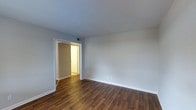 This single-floor space presents a clean and functional interior characterized by rich, dark hardwood-style flooring that runs throughout, adding warmth and visual continuity to the layout. The main room is finished with smooth, light grey walls and crisp white trim and baseboards, creating a neutral palette well-suited to a variety of furnishing styles. The ceiling is painted white, contributing to a bright and airy atmosphere. A doorway with white framing connects to an adjacent room, which is visible in the background and appears to continue the same flooring material. The connected space leads further into what appears to be an additional room or hallway, suggesting a thoughtfully linked floor plan. A ceiling vent is positioned near the doorway, indicating a central HVAC system for climate control. Electrical outlets are placed at standard heights along the walls. The overall condition of the space appears well-maintained, with fresh paint and clean finishes throughout. The open, unfurnished layout allows for a clear view of the space's proportions and flow, making it easy to envision a range of furniture arrangements and uses.