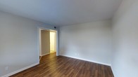 This single-floor space presents a clean and functional interior characterized by rich, dark hardwood-style flooring that runs throughout, adding warmth and visual continuity to the layout. The main room is finished with smooth, light grey walls and crisp white trim and baseboards, creating a neutral palette well-suited to a variety of furnishing styles. The ceiling is painted white, contributing to a bright and airy atmosphere. A doorway with white framing connects to an adjacent room, which is visible in the background and appears to continue the same flooring material. The connected space leads further into what appears to be an additional room or hallway, suggesting a thoughtfully linked floor plan. A ceiling vent is positioned near the doorway, indicating a central HVAC system for climate control. Electrical outlets are placed at standard heights along the walls. The overall condition of the space appears well-maintained, with fresh paint and clean finishes throughout. The open, unfurnished layout allows for a clear view of the space's proportions and flow, making it easy to envision a range of furniture arrangements and uses.